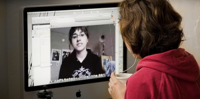 A woman sitting at an Apple computer looking at the screen, with her back to us. On the screen is a young woman that she is in a video call with.